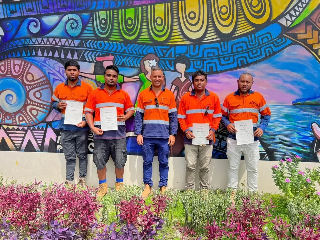 Five men in orange and blue work uniforms stand in front of a colorful mural, each holding a certificate. There are flowering plants in front of them.