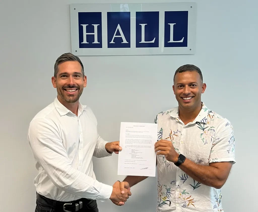 Two men stand in front of a "HALL" sign, smiling and shaking hands while holding a document together.