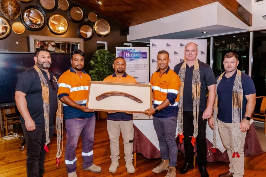 Six men pose indoors, five wearing orange and blue work uniforms with woven sashes, holding a framed boomerang; a display board and round wall decor are in the background.