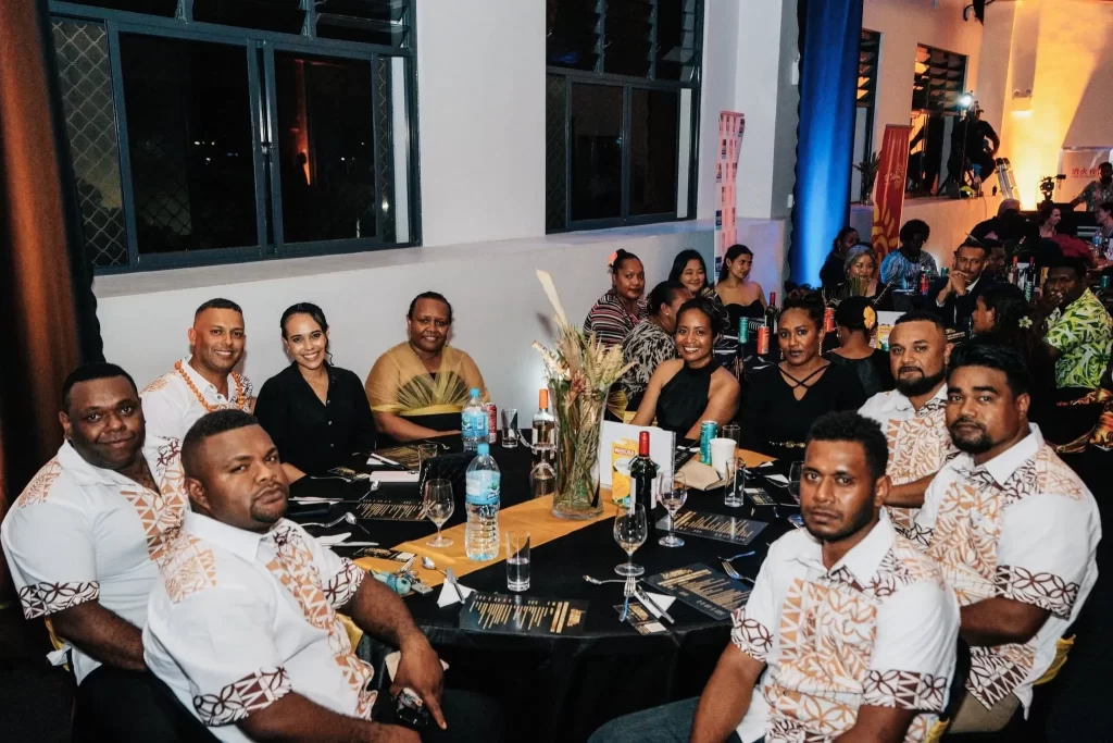 A group of people sit around a banquet table dressed in coordinated attire at a formal indoor event, with others seated at tables in the background.