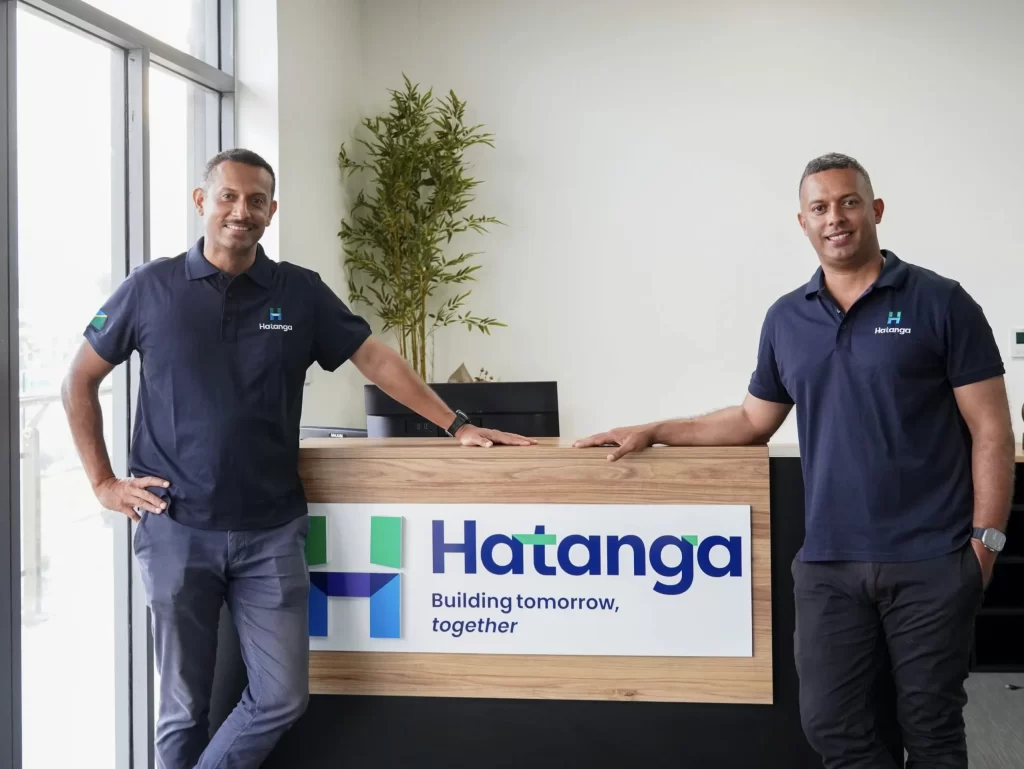 Two men in matching navy shirts stand by a reception desk with a "Hatanga" sign and the slogan "Building tomorrow, together" in an office setting.