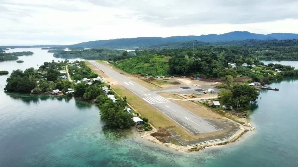 Aerial view of a narrow airport runway surrounded by water and greenery, with small buildings along the edge and forested hills in the background.