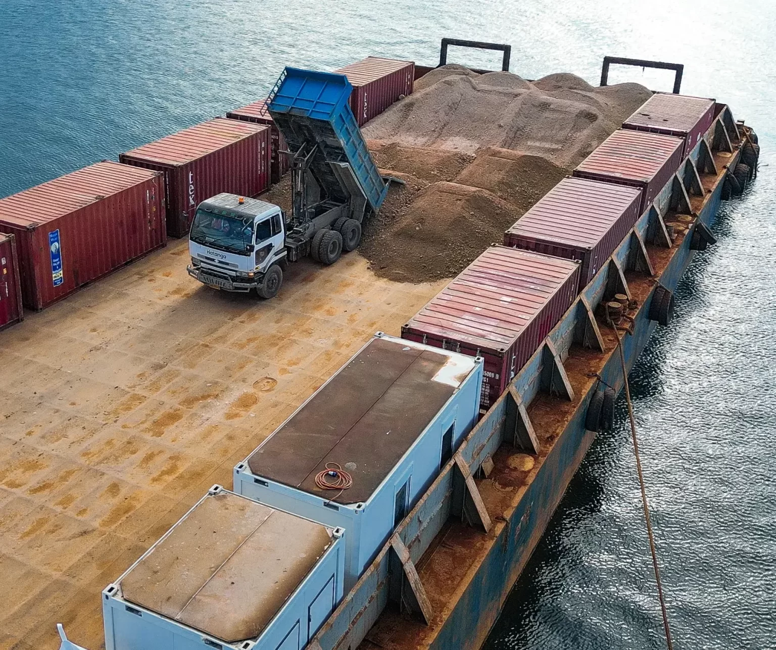 A dump truck unloads sand onto a large barge loaded with shipping containers, floating on calm water.
