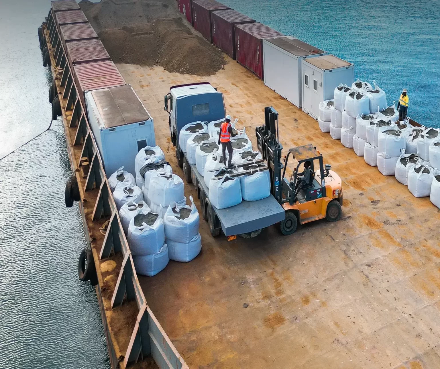 A forklift loads large white bulk bags onto a truck on the deck of a cargo ship, with containers, soil piles, and two workers in safety gear visible.