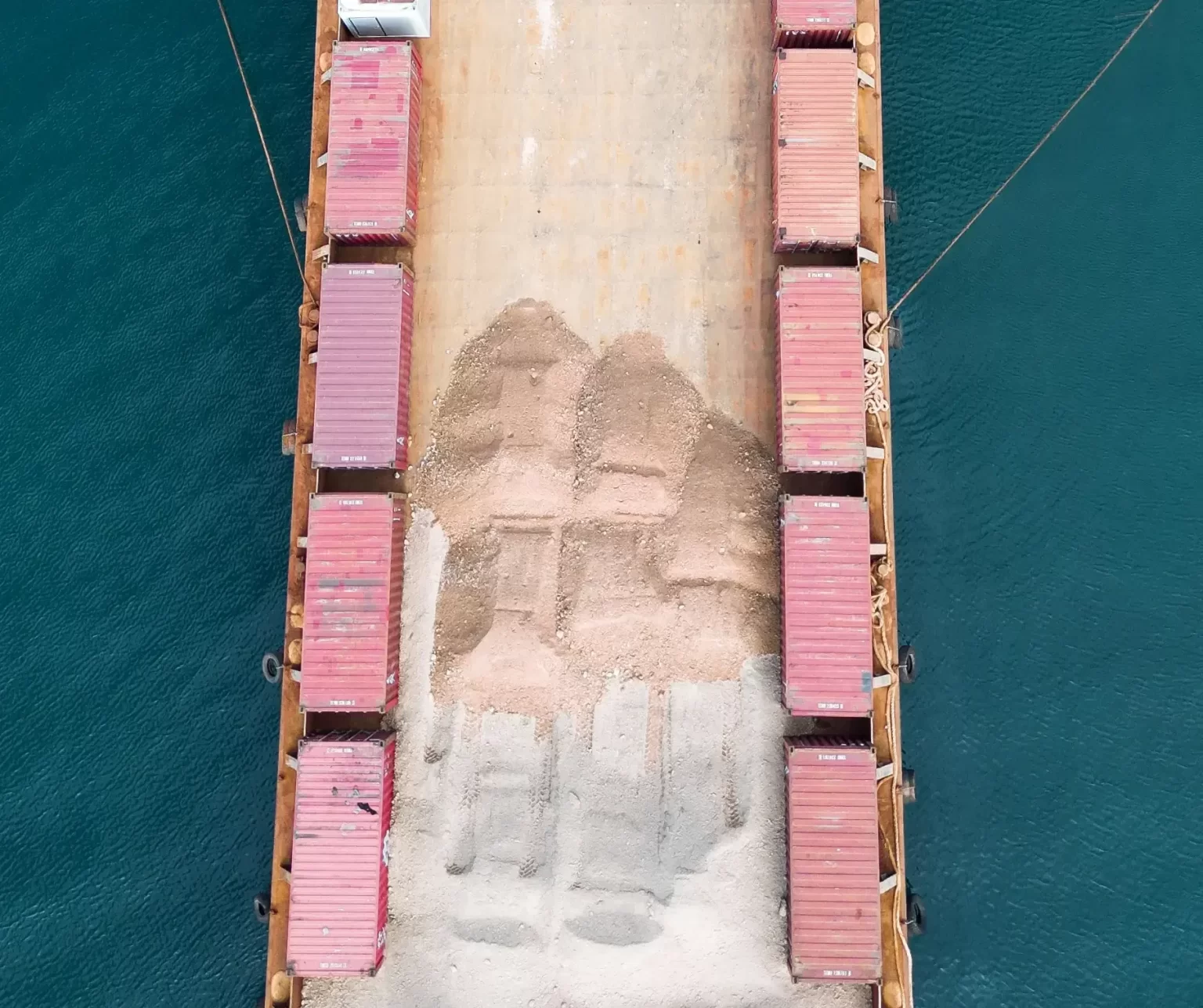 Aerial view of a cargo ship deck carrying red containers and large piles of loose granular material, possibly sand or gravel, surrounded by blue water.