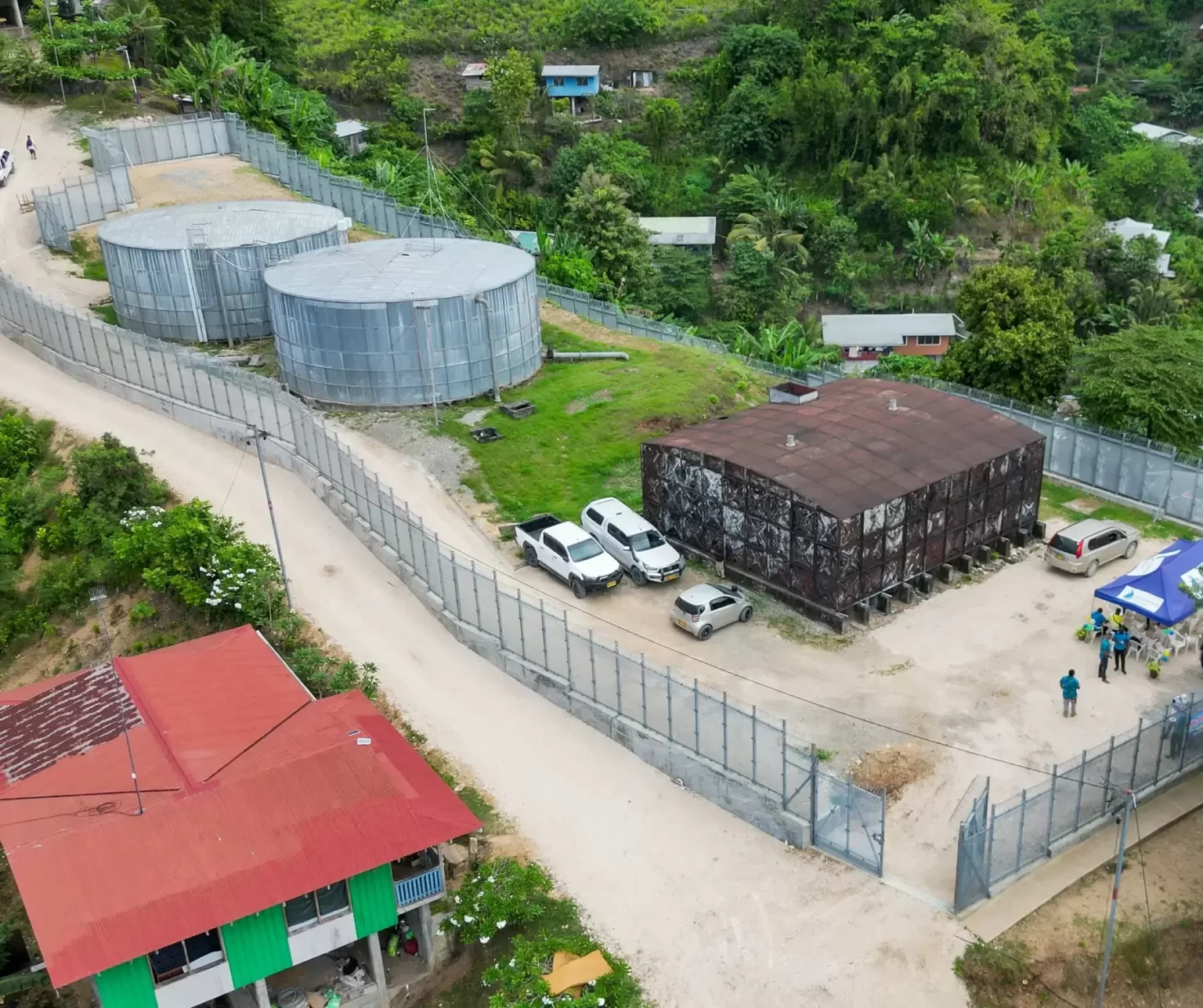 A fenced facility with two large cylindrical tanks, a rusted building, parked vehicles, and a few people standing outside, surrounded by greenery and scattered houses.