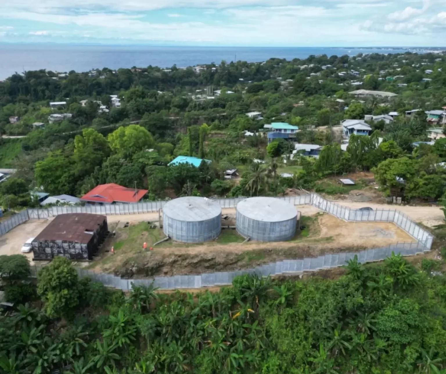 Aerial view of two large water storage tanks surrounded by a fence, with houses and dense greenery in the background near a coastline.