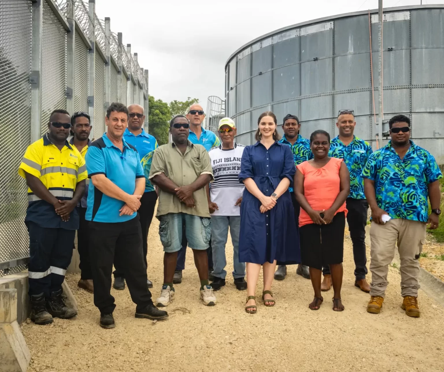 A group of twelve people stand outdoors on a gravel path near a chain-link fence and a large metal tank, posing for a group photo.