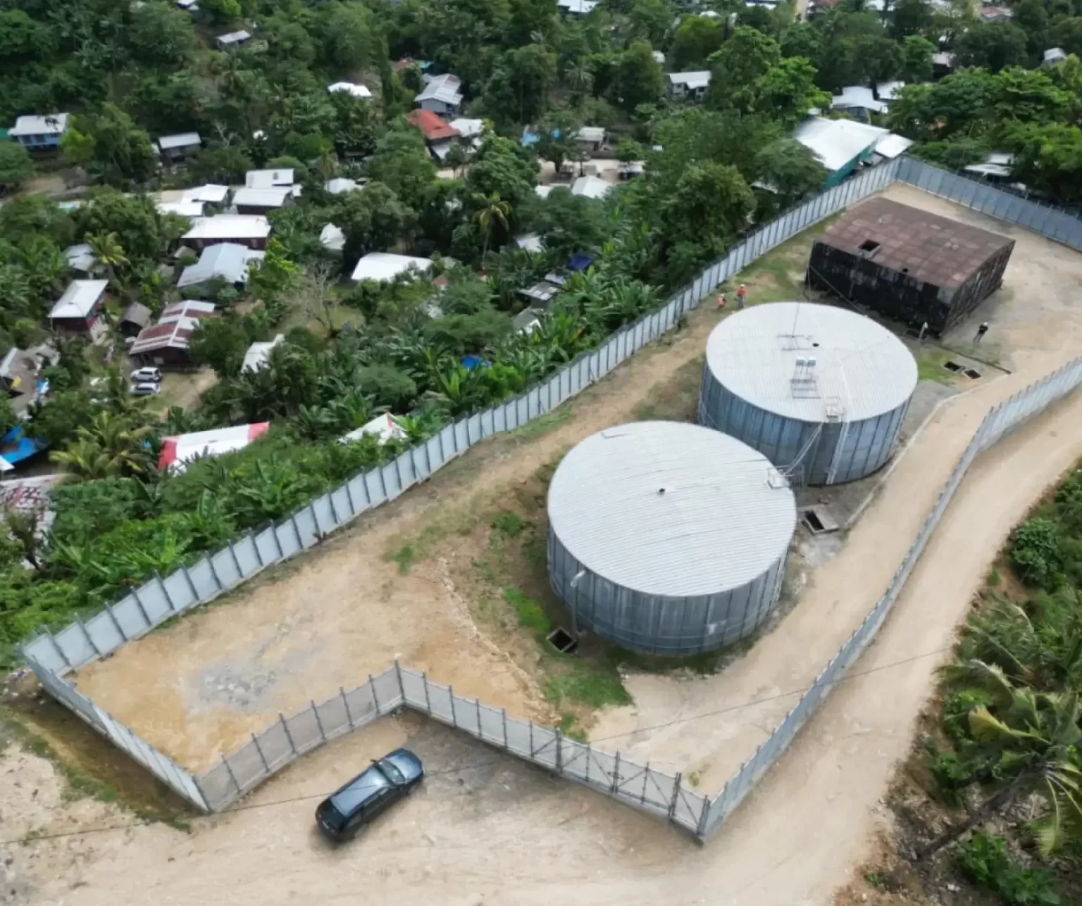 Aerial view of a fenced facility with two large circular storage tanks and a rectangular building, located near a dirt road and surrounded by trees and residential houses.