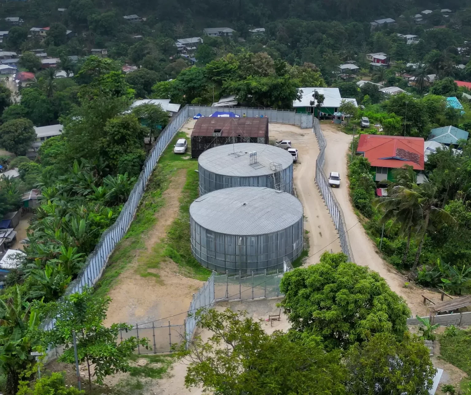 Aerial view of two large cylindrical fenced water tanks inside a secured facility, surrounded by trees and nearby residential buildings.