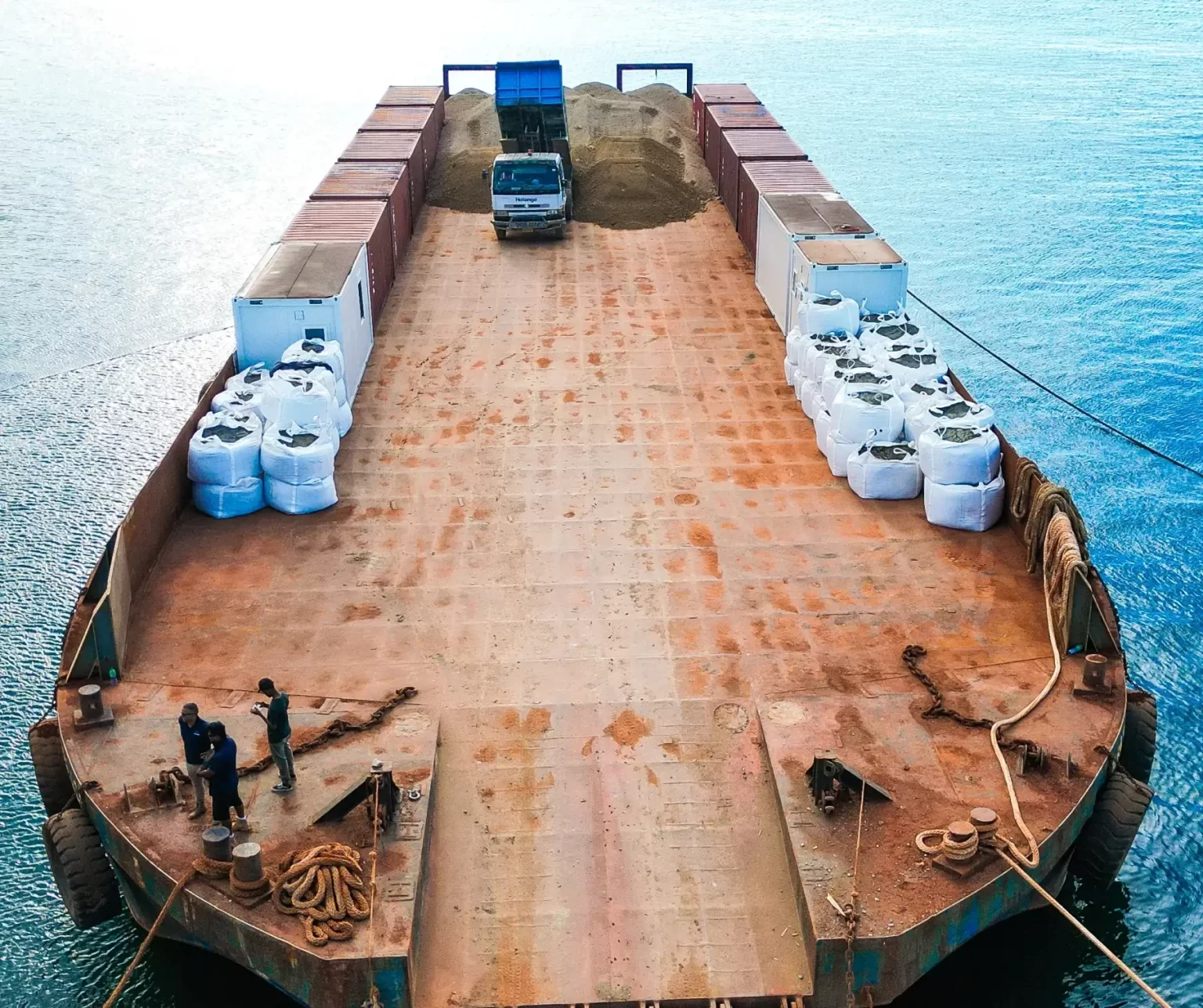 A truck unloads sand onto a large, rusted cargo barge carrying white bulk bags, while two workers stand near ropes at the bow on calm water.