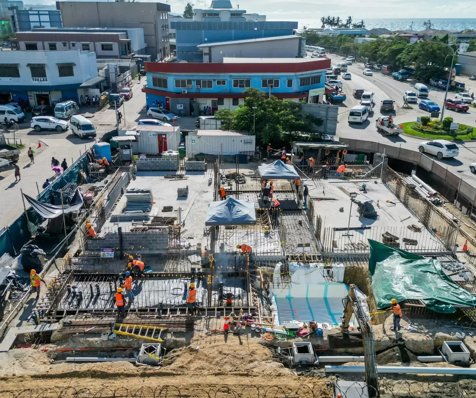 Aerial view of a construction site with workers in orange safety vests building the foundation of a multi-story structure, surrounded by buildings and parked vehicles.