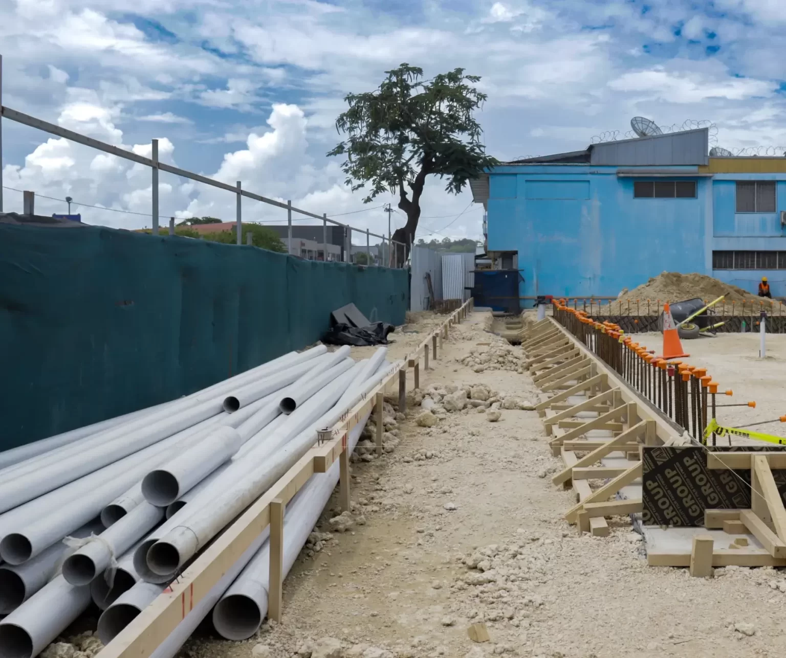 Construction site with stacked PVC pipes on the left, wooden formwork and caution tape along a dirt path, and a blue building in the background.