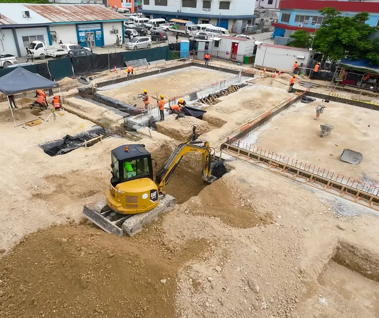 A construction site with workers and a yellow excavator digging and moving soil around laid foundation structures.