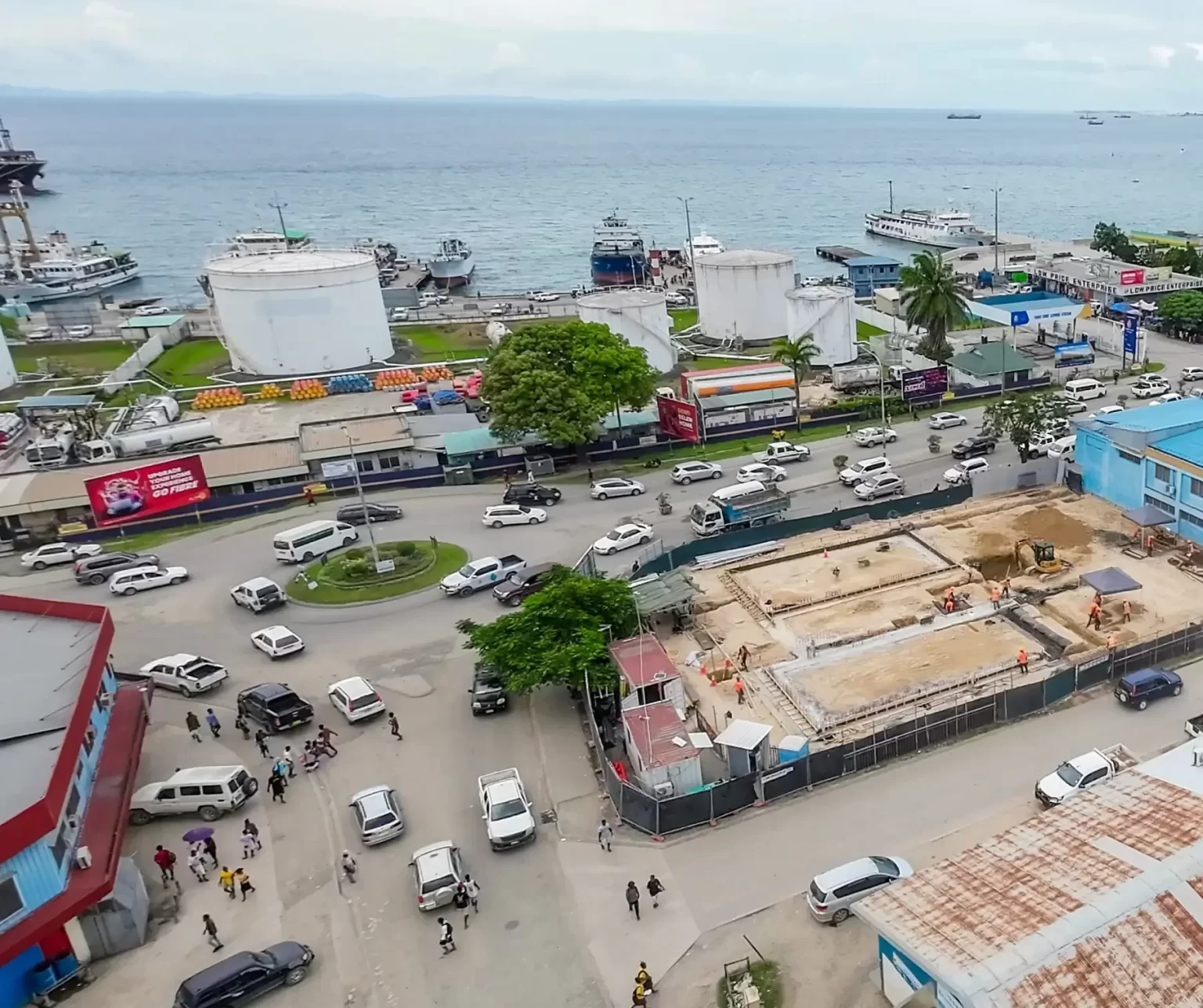 Aerial view of a busy coastal city with traffic, construction site, commercial buildings, and ships docked near large storage tanks by the waterfront.