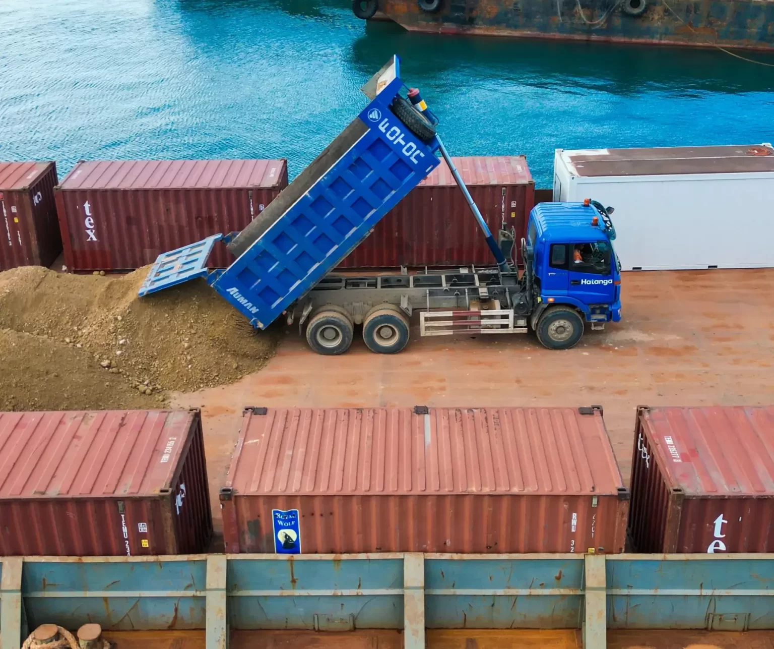 A blue dump truck unloads a pile of dirt onto a dock, surrounded by stacked shipping containers near the water.