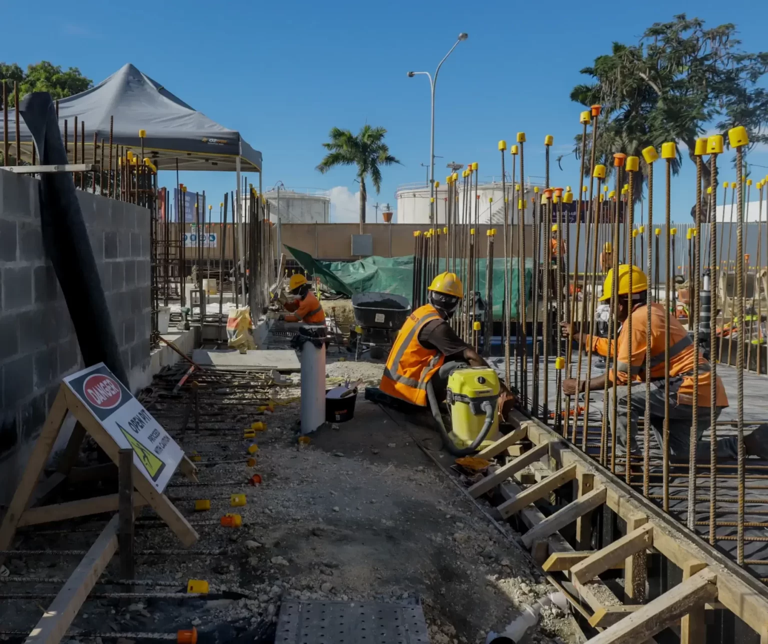Construction workers in safety gear install rebar and work on the foundation of a building site under clear blue skies, with tools and materials scattered around.