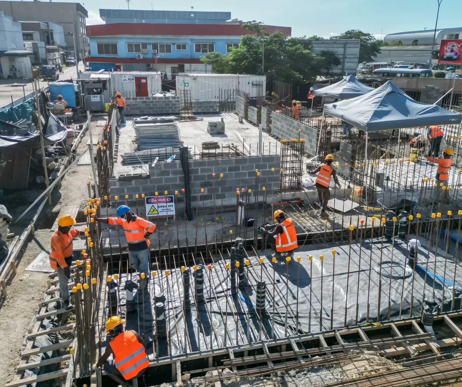 Workers in safety gear construct a building foundation with rebar and concrete blocks at an outdoor construction site during daytime.