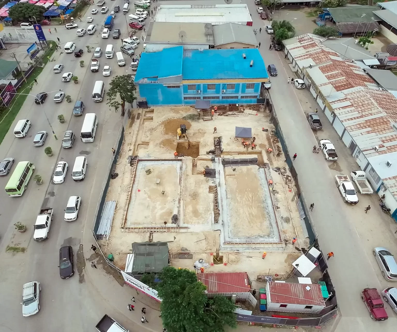Aerial view of a construction site with building foundations visible, surrounded by a fence, adjacent to a busy street and a blue-roofed building.