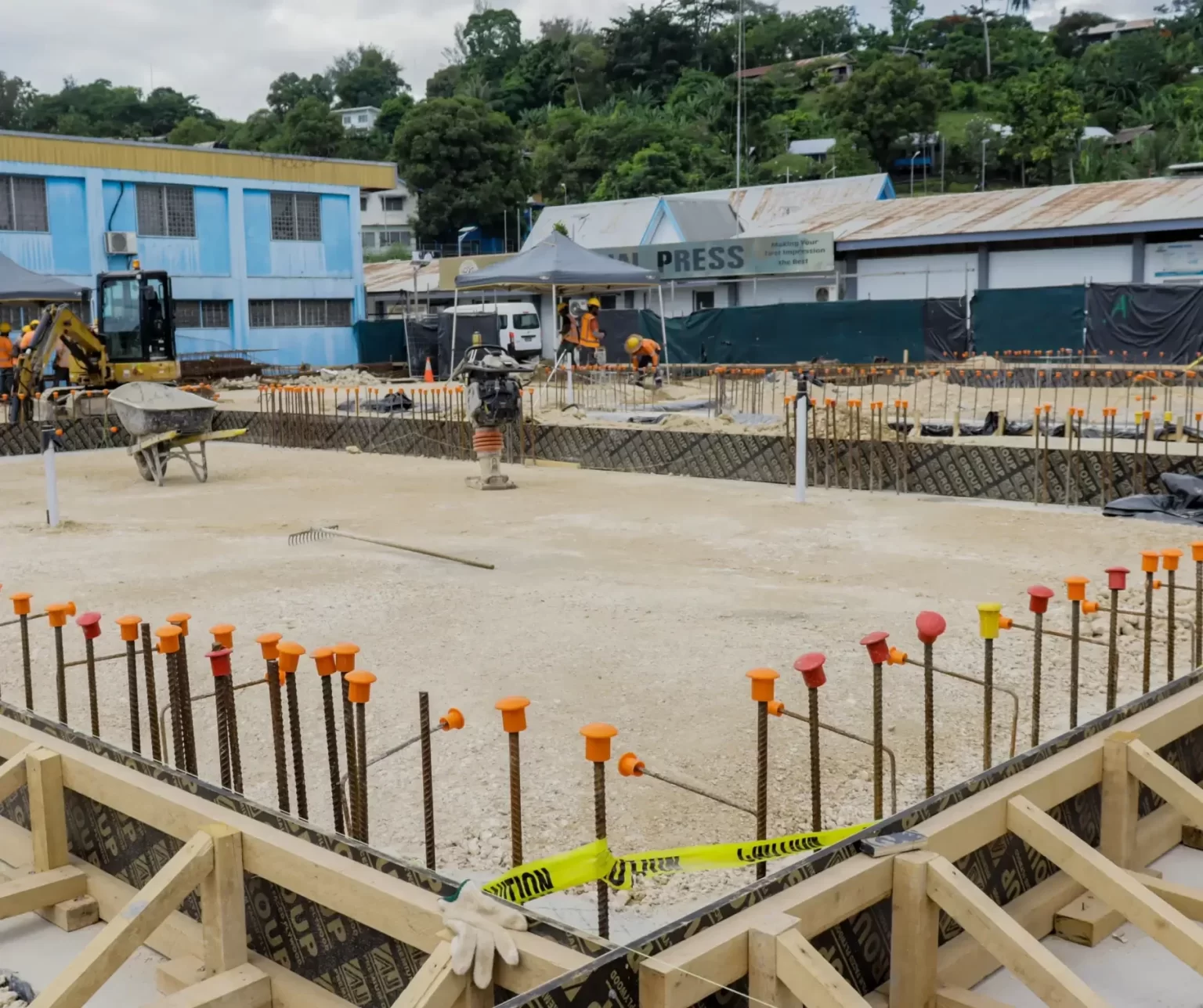 Construction site with exposed rebar, warning caps, caution tape, and equipment visible; workers and buildings are in the background.