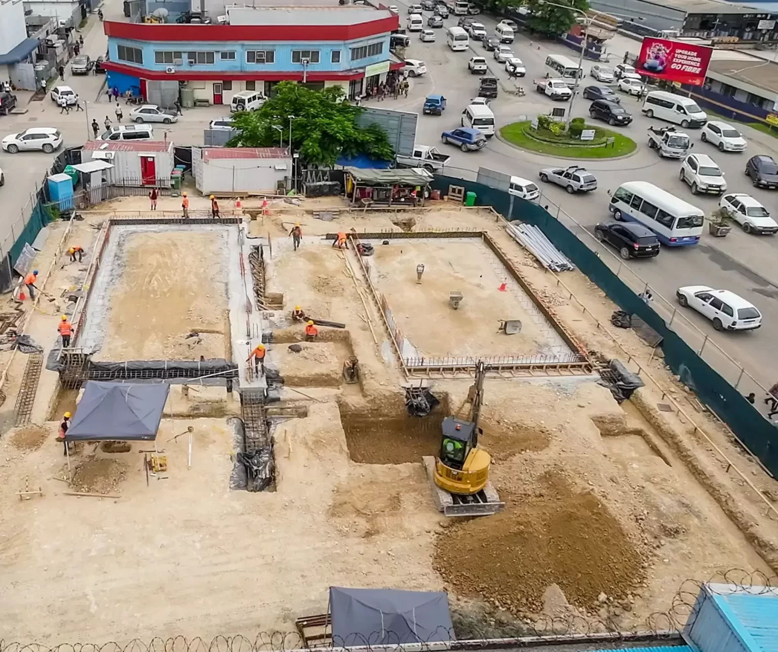 Aerial view of a construction site with workers, machinery, and building foundations, surrounded by busy roads and traffic in an urban area.
