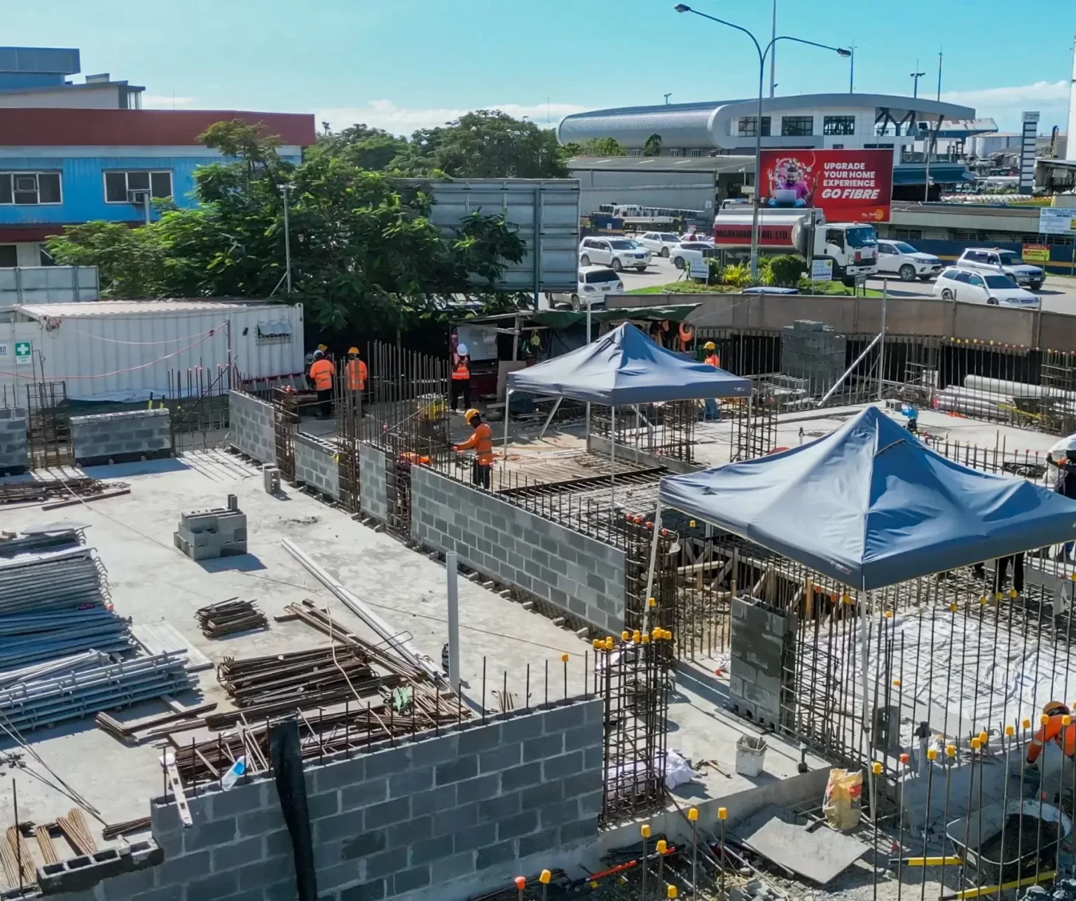 Construction site with workers building concrete block walls and metal rebar, two blue tents for shade, and traffic moving on a road with buildings and billboards in the background.