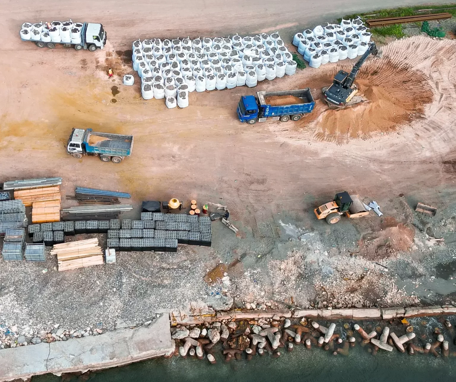 Aerial view of a construction site with trucks, stacked materials, large white bags, an excavator, and debris near a waterfront.
