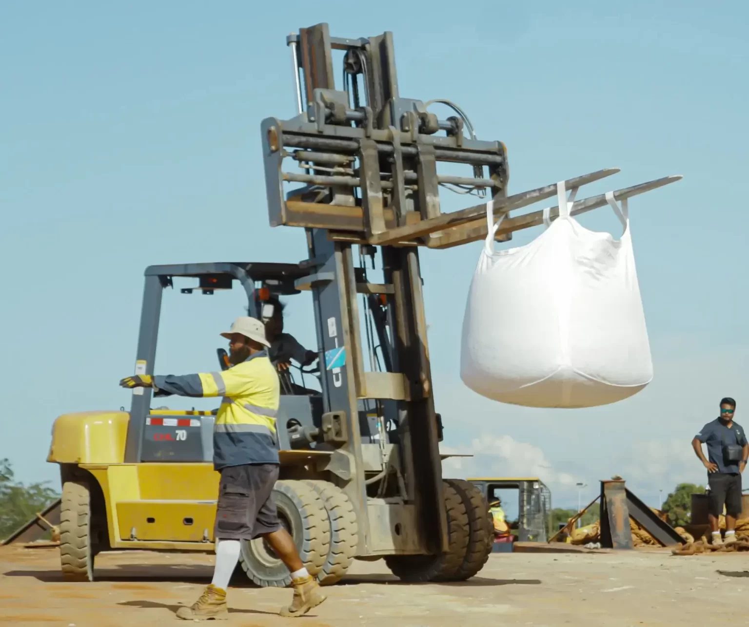 A worker operates a forklift carrying a large white bulk bag at a construction site, while another person stands in the background.