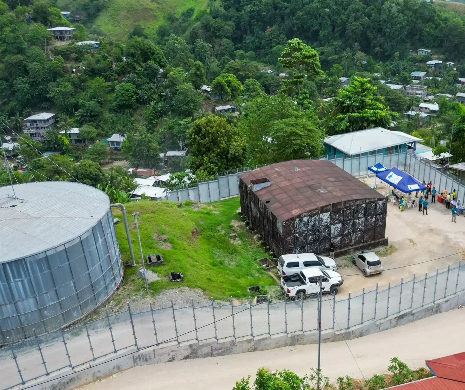 A fenced compound with metal buildings, parked cars, and people gathered under a canopy, surrounded by greenery and houses on a hillside.