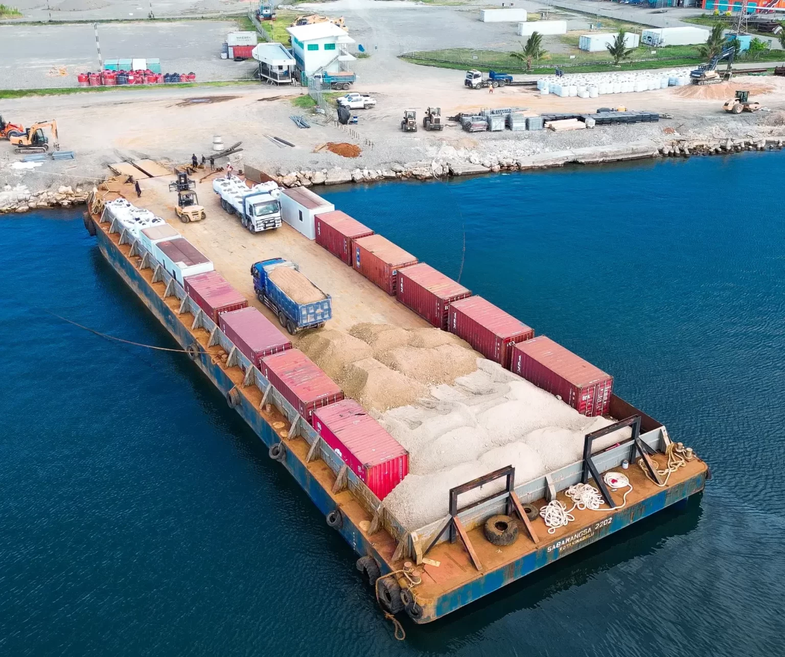 A barge loaded with shipping containers and sand is docked at an industrial waterfront area with various vehicles and equipment nearby.
