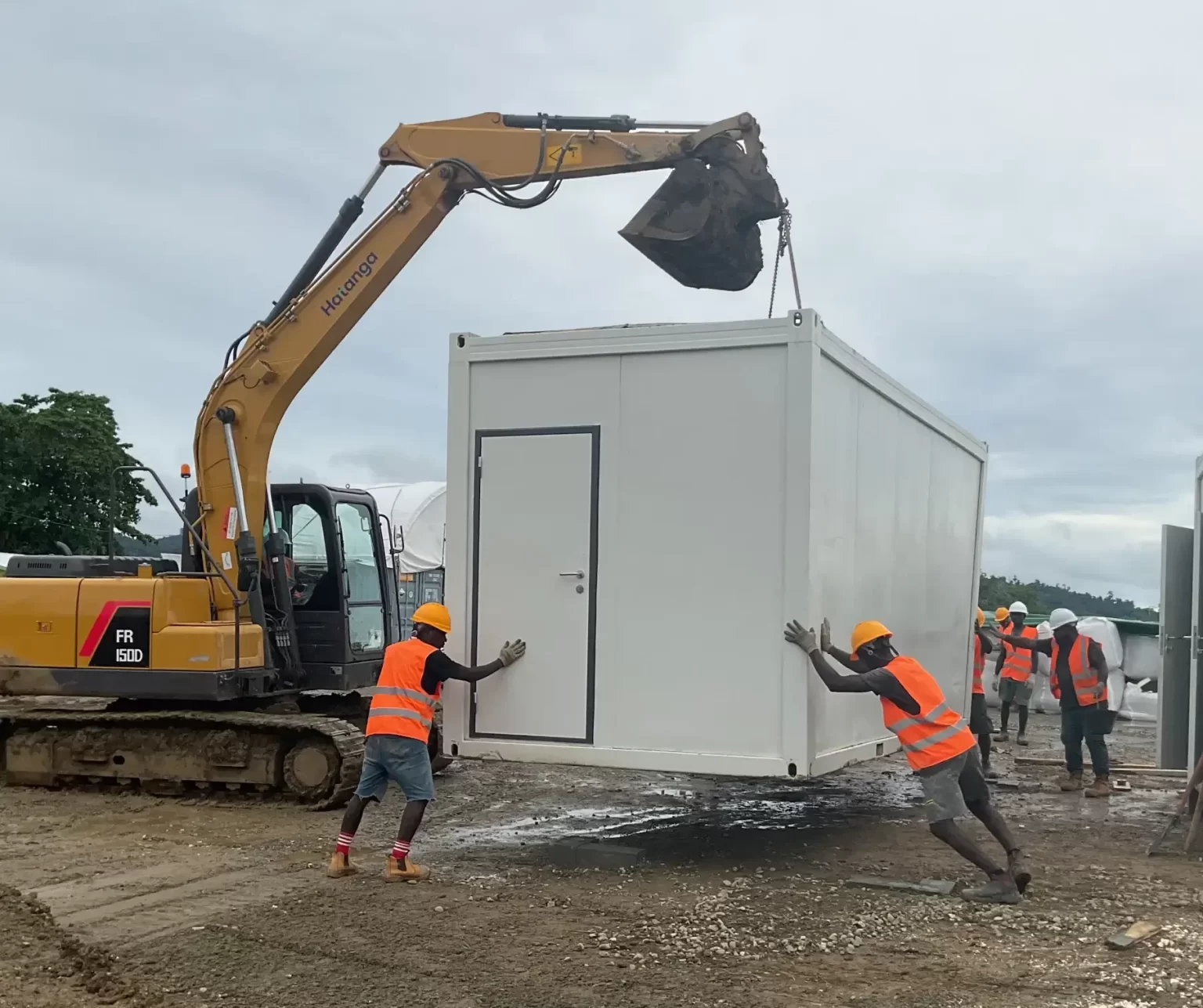 Construction workers in orange vests guide a prefabricated building module being lifted by an excavator on a muddy construction site.