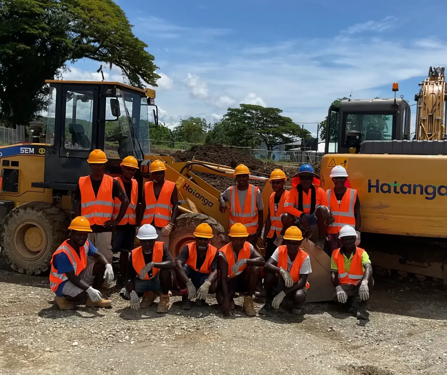 A group of construction workers in orange safety vests and hard hats pose in front of heavy machinery at a construction site on a sunny day.