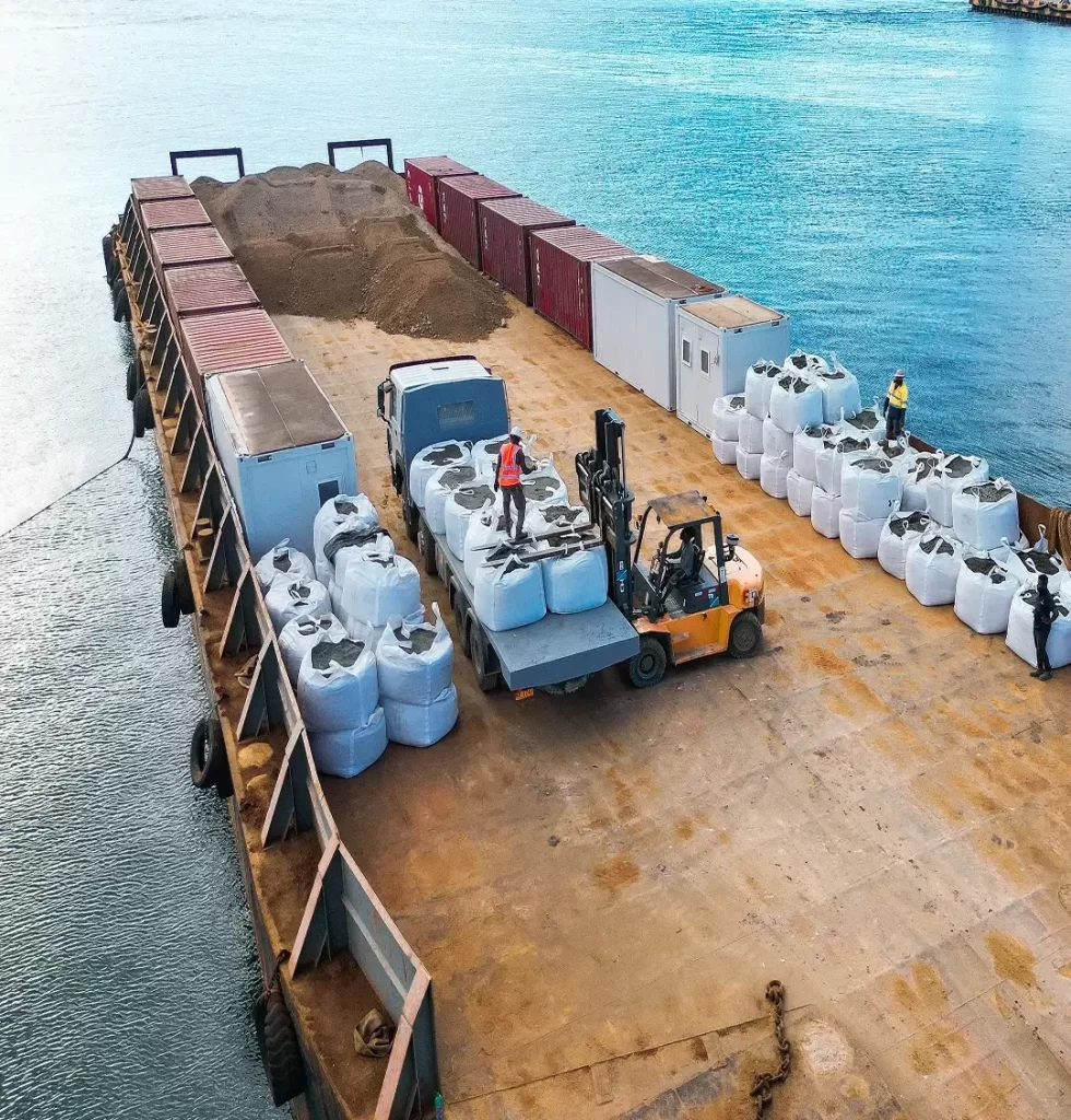 A forklift loads large white bags onto a truck on a cargo barge, with additional containers, bags, and a pile of soil nearby. Two workers in safety gear are present.