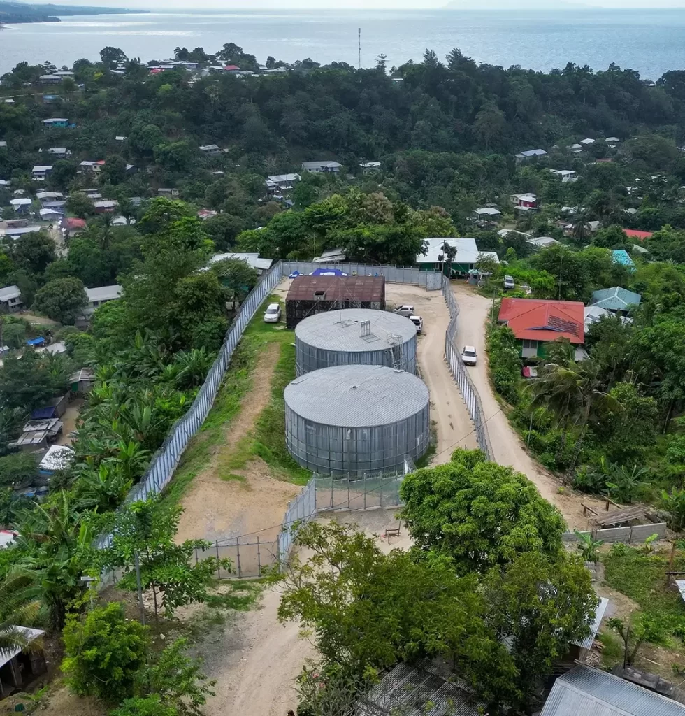 Aerial view of two large cylindrical water tanks enclosed by a fence, with surrounding trees, houses, and a coastline in the background.